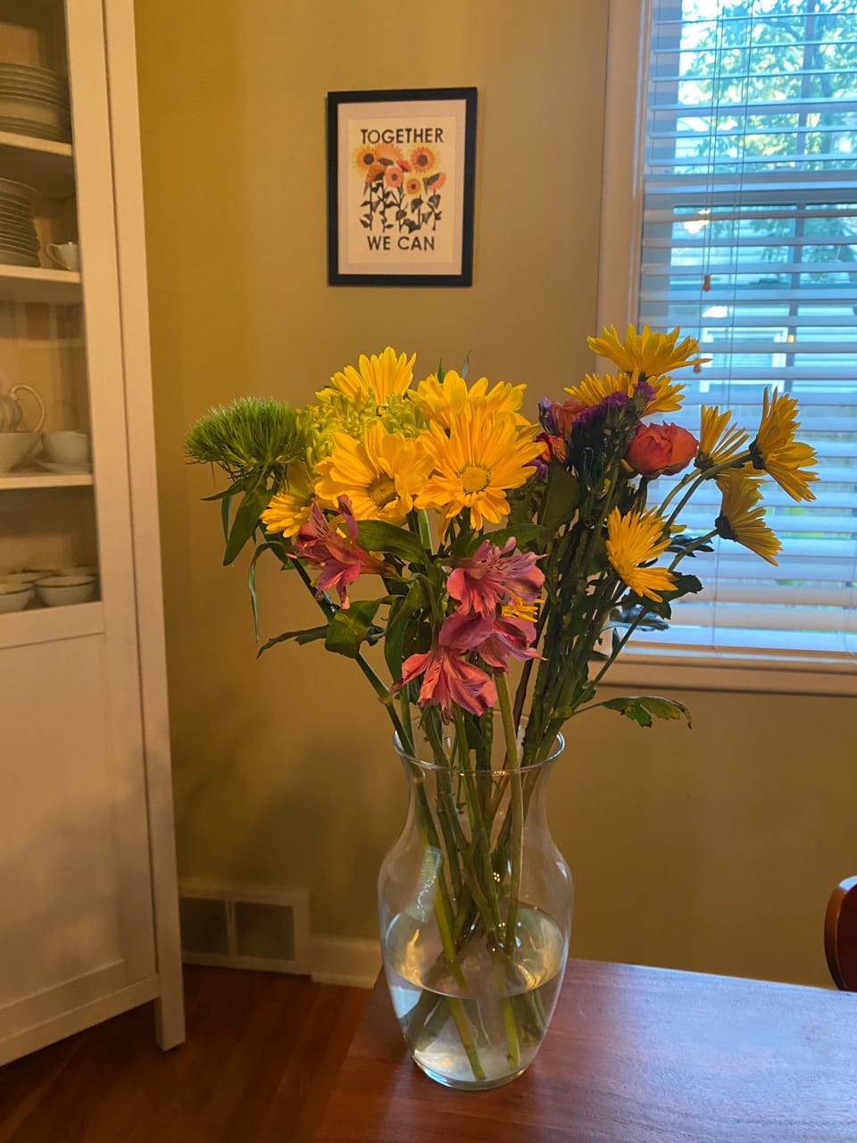 A vase of flowers sits on a table in front of a picture of similar looking flowers reading "Together We Can."